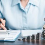 A person sitting at a desk managing personal finances, dropping a coin into a clear glass jar filled with money next to stacks of coins arranged in increasing height, while using a smartphone or calculator placed on financial documents.