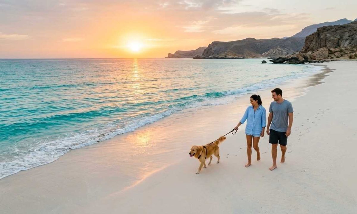 A young couple walking their golden dog along a serene, white sand beach with turquoise water and Omani coastline cliffs under a warm sunrise sky.