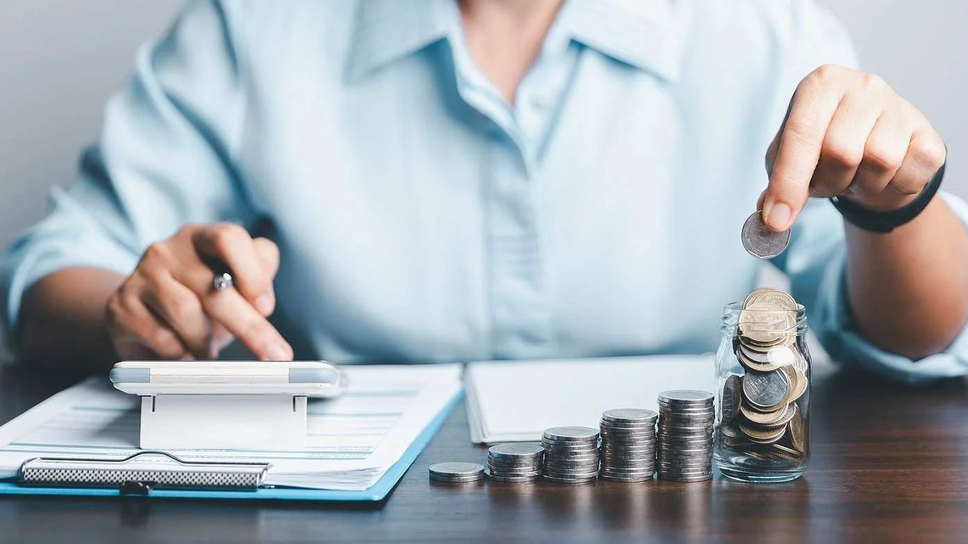 A person sitting at a desk managing personal finances, dropping a coin into a clear glass jar filled with money next to stacks of coins arranged in increasing height, while using a smartphone or calculator placed on financial documents.