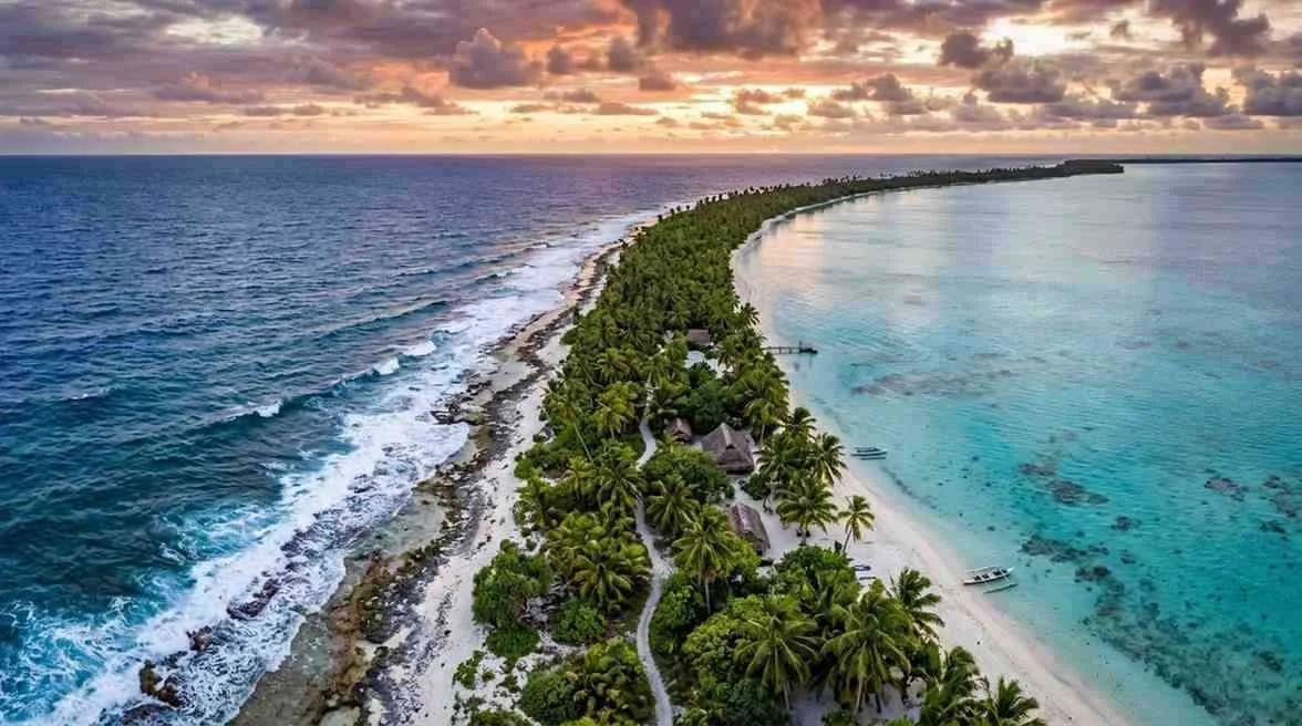 Aerial view of a thin strip of lush palm trees on a low-lying coral atoll separating deep blue ocean waves from a calm turquoise lagoon.